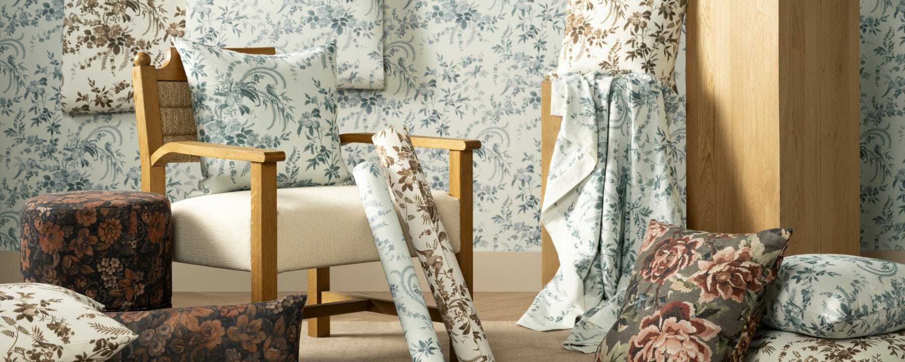A cozy room with a wooden armchair, floral cushions, fabric rolls, and pillows in blue, brown, and rose patterns, set against a floral wallpaper background in soft blue and white tones, by McGee & Co.