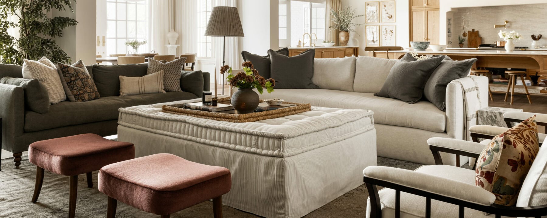A stylish living room with a large beige sectional sofa, gray sofa, white armchairs, a tufted ottoman coffee table, red stools, decorative pillows, and plants, with natural light streaming through tall windows, by McGee & Co.